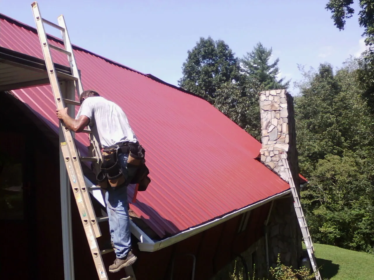Roofer on a ladder beside a red metal roof and stone chimney — Soffit & Fascia Repair by Roofing Brothers in Lawtell